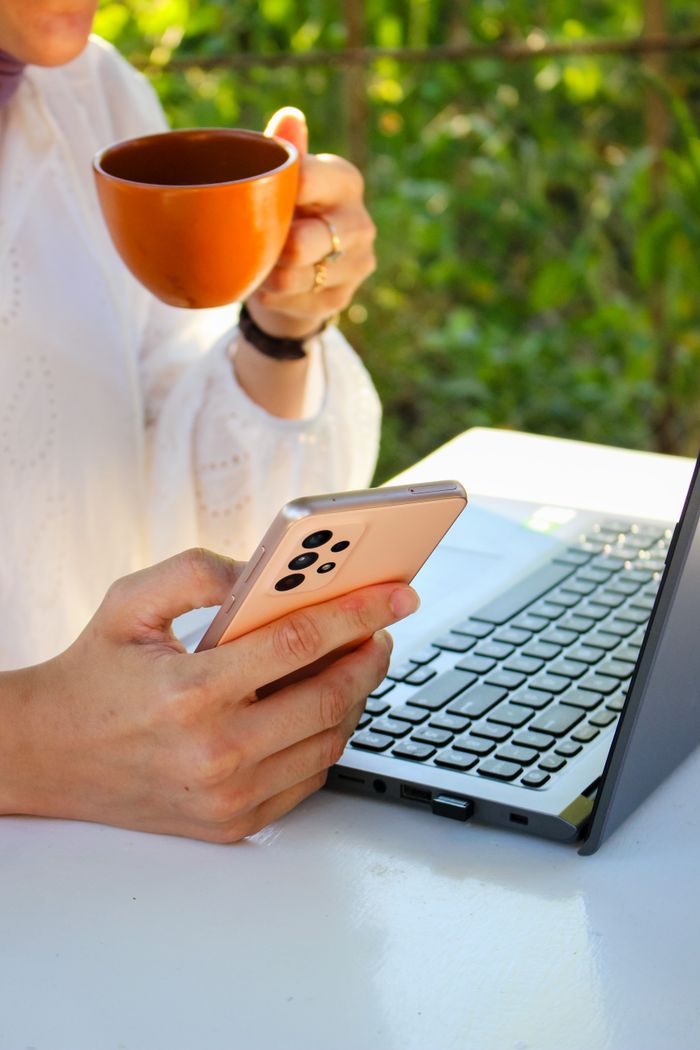 Person using a smartphone while drinking coffee near a laptop outdoors, showcasing multitasking with technology.